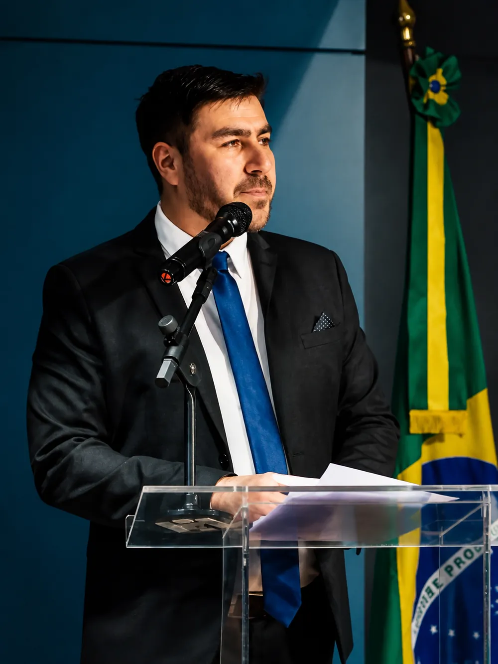 Fernando Zanuzo conduzindo uma cerimônia ao microfone, ao lado da bandeira do Brasil