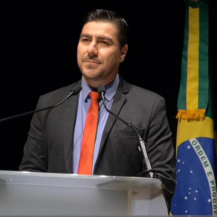 Fernando Zanuzo de terno e gravata ao púlpito, diante do público, com a bandeira do Brasil ao fundo